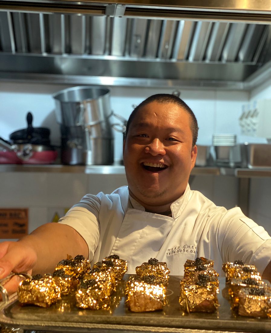 Chef Trần Quốc Đạt smiling with a tray of gold-leaf canapés